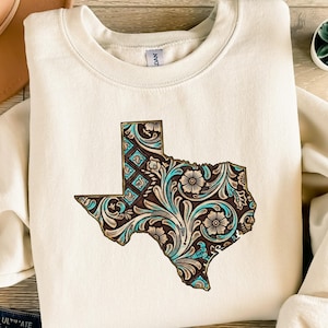 Könnte beinhalten: Cremefarbenes Sweatshirt mit einer detaillierten Grafik des Bundesstaates Texas. Die Texas-Umrisse sind mit einem braunen, türkisfarbenen und cremefarbenen Blumenmuster gefüllt. Das Sweatshirt hat einen Rundhalsausschnitt und lange Ärmel.