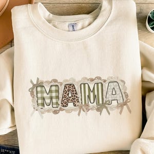 Könnte beinhalten: Cremefarbenes Sweatshirt mit dem Wort "MAMA" in einem dekorativen Design. Die Buchstaben zeigen Karomuster, Leopardenmuster und florale Muster, umrahmt von einem spitzenartigen Rand und kleinen Schleifen. Ein gemütliches und stilvolles Oberteil.