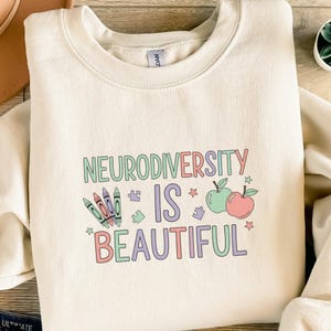 Könnte beinhalten: Cremefarbenes Sweatshirt mit den Worten "NEURODIVERSITY IS BEAUTIFUL" in bunten Buchstaben. Das Design umfasst Buntstifte, Puzzleteile, Äpfel und Sterne. Ein bequemes, lässiges Oberteil.
