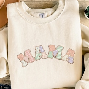 Könnte beinhalten: Cremefarbenes Sweatshirt mit dem Wort "MAMA" in einem Patchwork-Design auf der Vorderseite. Die Buchstaben sind in Pastellfarben wie Rosa, Grün und Lila gehalten, mit Punkten und floralen Mustern.