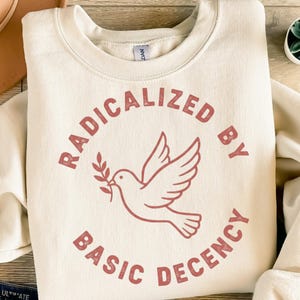 Könnte beinhalten: Cremefarbenes Sweatshirt mit dem Text "RADICALIZED BY BASIC DECENCY" in einem kreisförmigen Design. Eine rote Taube mit einem Olivenzweig befindet sich in der Mitte. Das Sweatshirt hat eine neutrale Farbe.