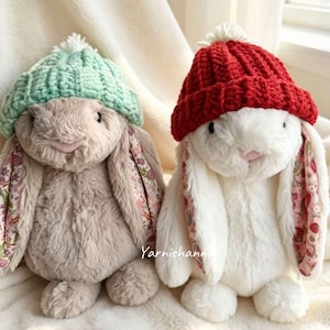 May include: Two plush stuffed rabbits with long ears featuring floral fabric linings. The left rabbit is light brown with a mint green knit beanie. The right rabbit is white with a red knit beanie. The text Yarniehannah is visible in the center.