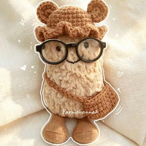 May include: A plush tan crochet doll with a fuzzy texture, wearing a matching tan crochet hat with bear ears and a small crossbody bag. The doll features black round glasses, a stitched smile, and tan feet. The text Yarniehannah is visible.
