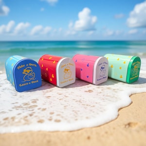 Puede incluir: Cuatro buzones metálicos de colores, azul, rojo, rosa y verde, están alineados en una playa de arena con el océano al fondo. Cada buzón tiene una parte delantera blanca con un dibujo de pato y el texto "Take a Duck, Leave a Duck".