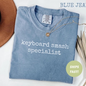 May include: A blue jean colored t-shirt with the words "keyboard smash specialist" printed in white. The shirt has a crew neck and short sleeves. A gold necklace with a pendant is draped on the shirt.