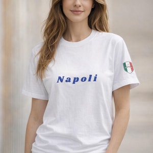 Può includere: T-shirt bianco con la scritta "Napoli" in blu sul petto. Un piccolo stemma con la bandiera italiana è sulla manica sinistra. La maglietta è di stile casual.
