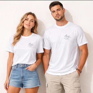 Può includere: Due persone indossano magliette bianche con la parola "Asanje" stampata in un carattere corsivo sulla parte superiore sinistra del petto. Le magliette hanno maniche corte e girocollo. La donna indossa pantaloncini di jeans e l'uomo pantaloncini color cachi.