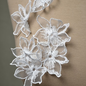 May include: White floral lace applique featuring intricate embroidered petals with sheer mesh centers. The delicate white threadwork forms layered flower shapes with defined outlines, secured with small silver pins to a tan fabric surface.