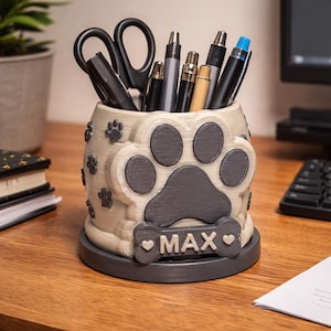Puede incluir: Un portalápices beige y gris con huellas de patas con el nombre "MAX" en una placa con forma de hueso. El portalápices está lleno de bolígrafos, un rotulador y unas tijeras. El portalápices está sobre un escritorio de madera.