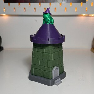 May include: A 3D-printed green and purple castle tower with a green frog wearing a purple witch hat on top. The tower is green with a stone block design, a purple roof, and a small door.