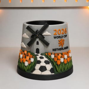 Puede incluir: Una maceta cilíndrica gris con un diseño en relieve de un molino de viento negro, tulipanes naranjas y blancos, y un camino con patrón de balón de fútbol. El texto 2026 WORLD CUP NETHERLANDS y un león naranja están grabados en el lateral.