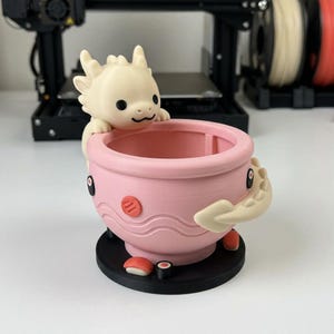 May include: A 3D-printed pink planter pot with a dragon design. The dragon is cream-colored and peeking over the edge of the pot. The pot has sushi-shaped details and a black base. A unique decorative item.