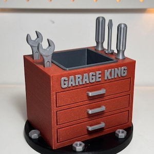 May include: A red miniature toolbox with the text "GARAGE KING" on the front. The toolbox has three drawers, gray wrenches, screwdrivers, and nuts. The toolbox sits on a black circular base.