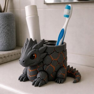 May include: A gray dragon-shaped toothbrush holder with orange accents. The holder has multiple compartments for toothbrushes and toothpaste. A white tube of toothpaste and a blue and white toothbrush are visible.