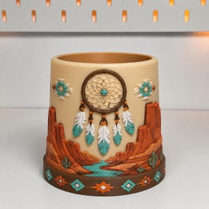 May include: A cylindrical beige planter featuring a raised relief design of a desert landscape with red rock formations, a blue river, and green cacti. A central dreamcatcher motif with white and teal feathers is displayed above the landscape.