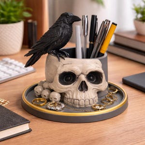 May include: A decorative desk organizer featuring a skull-shaped pen holder with a black raven perched on top. The organizer includes a tray with gold-colored accents and small skull details, holding pens and jewelry.