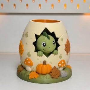 Può includere: Un vaso a forma di uovo color crema con un dinosauro verde che sbuca da un'apertura a forma di stella. L'esterno è decorato con zucche arancioni, foglie autunnali marroni e arancioni e funghi arancioni a pois bianchi su una base verde.