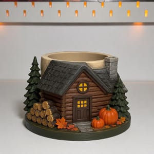 May include: A cylindrical ceramic planter shaped like a log cabin with a dark grey roof, a stone chimney, and a brown door. The exterior features two green pine trees, a stack of round logs, two orange pumpkins, and scattered autumn leaves.