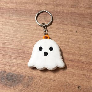 May include: A white ghost-shaped keychain with black eyes and mouth, attached to a silver keyring. The ghost has a wavy bottom edge and an orange accent. The keychain is a Halloween-themed accessory.