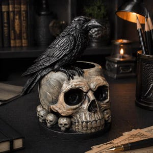 Op de afbeelding: Een zwart ravenbeeldje bovenop een schedelvormige container. De container is gedetailleerd met een schedelgezicht en kleinere schedels rond de basis. De raaf heeft gedetailleerde veren en een prominente snavel. De algehele esthetiek is gotisch.