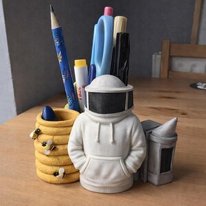 May include: A novelty desk tidy with a bee-keeping theme. It features a white hooded figure wearing a beekeeper's helmet, a honeycomb-shaped pencil pot with bee details, and a small beehive. Various pens and pencils are visible.