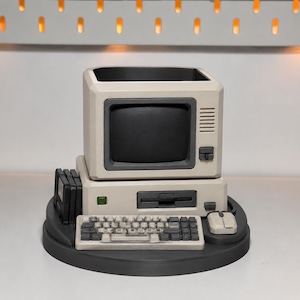 Könnte beinhalten: Ein beigefarbener und dunkelgrauer Schreibtisch-Organizer in Form eines Vintage-Personal-Computers. Das Objekt verfügt über einen hohlen Monitorbildschirm zur Aufbewahrung, eine Tastatur, eine Maus und drei kleine Disketten an der Seite, montiert auf einem runden Sockel.
