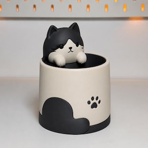 Könnte beinhalten: Ein zylindrischer Schreibtisch-Organizer mit einer schwarz-weißen Katzenfigur, die über den Rand schaut. Der Behälter ist cremeweiß mit einem schwarzen Boden und einer schwarzen Pfotenabdruck-Grafik an der Seite. Die Katze hat schwarze Ohren, ein weißes Gesicht und schwarze Augen.