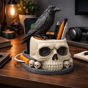 May include: A desk organizer shaped like a skull with a black raven perched on top. The organizer has a compartment for pens and a slot for a phone. The base is gray with small skull accents. The raven and skull are detailed.