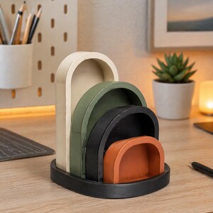 May include: A multi-colored desk organizer with an arched design. The organizer features a base and four nested compartments in cream, green, black, and orange. The organizer is made of wood and sits on a wooden desk.