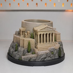 Op de afbeelding: Een ronde plantenbak met een gedetailleerd 3D-geprint model van de Akropolis van Athene. De structuur bevat het Parthenon met zuilen, stenen muren en kleine groene cipressen op een getextureerde grijze rotsachtige ondergrond.