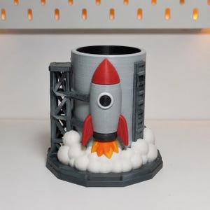 以下が含まれることがあります： 3Dプリントされたロケットデザインの円筒形デスクオーガナイザー。ロケットはグレーで、赤いノーズコーンとフィンがあり、ベース部分の白い雲に向かってオレンジ色の炎を噴射しています。側面にダークグレーの打ち上げタワーとハシゴが取り付けられています。