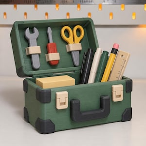 May include: A green plastic toolbox with a black handle and corner accents, featuring two beige latches. The open lid holds a gray wrench, a red and gray screwdriver, and yellow scissors. Inside are a beige block, pens, and a wooden ruler.