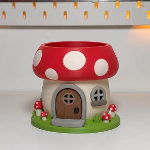 May include: A mushroom-shaped planter with a red cap featuring white spots. The base is cream-coloured with a brown arched door, a small window, and a green circular platform decorated with tiny red and white mushrooms and green grass blades.