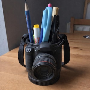 May include: Black desk organizer shaped like a camera, holding pens, pencils, and scissors. The organizer has a lens and strap detail. A blue pencil with a cartoon character is visible.
