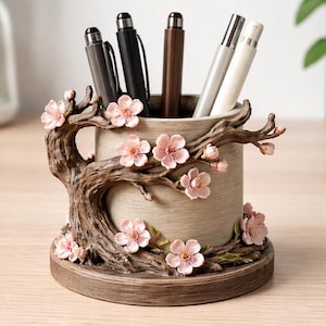 Puede incluir: Un portalápices decorativo beige con base marrón, adornado con una rama de árbol y flores de cerezo rosas. Contiene varios bolígrafos. Un accesorio de escritorio único.