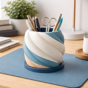 Puede incluir: Un organizador de escritorio decorativo con un diseño en espiral en tonos azul, blanco y melocotón. Contiene bolígrafos, lápices y tijeras. Se encuentra sobre una base azul a juego y una alfombrilla de escritorio azul, de unos 10 cm de altura.