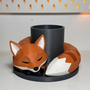 May include: A 3D-printed orange fox pen holder with a sleeping fox design. The fox has white accents on its ears and tail, black paws, and closed eyes. The pen holder is a dark grey cylinder.