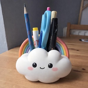 Puede incluir: Un organizador de escritorio blanco en forma de nube con una cara sonriente y mejillas rosadas. Un arco iris está detrás de la nube, sosteniendo bolígrafos, lápices y tijeras. El lápiz tiene un diseño azul.