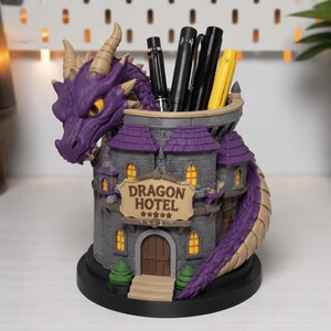 May include: A purple dragon head attached to a gray castle-shaped pen holder with a sign that reads DRAGON HOTEL and five stars. The structure features a brown door, small yellow-lit windows, and a long purple tail wrapping around the base.