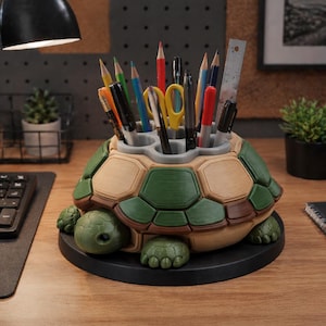 Op de afbeelding: Een schildpadvormige bureau-organizer in groen, beige en bruin, met diverse pennen, potloden en een liniaal. De organizer staat op een zwarte basis en heeft een gedetailleerd schildontwerp. De kop en poten van de schildpad zijn zichtbaar.