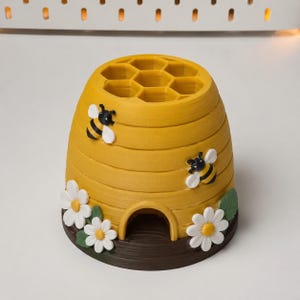 May include: A yellow beehive-shaped desk organizer with a honeycomb top featuring seven circular compartments. The exterior is decorated with two black and yellow bees, white daisy flowers with yellow centers, and green leaves on a dark brown base.