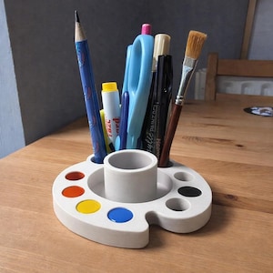 May include: A white, palette-shaped desk organizer holds various art supplies, including a blue pencil, scissors, markers, and a paintbrush. The palette has several colored paint wells in red, orange, yellow, and blue.