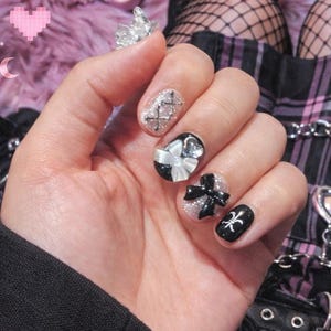 Puede incluir: Primer plano de una mano con uñas decoradas con purpurina plateada, lazos negros y blancos, sobre un fondo rosa y esponjoso. También se ven zapatos negros con detalles de cadena, un bolso plateado en forma de corazón y un teléfono móvil rosa.