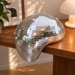 May include: A silver disco ball sculpture with a fluid, melting shape that drapes over the edge of a wooden surface. The object is covered in small, square, reflective mirror tiles that catch and scatter light across the surrounding area.