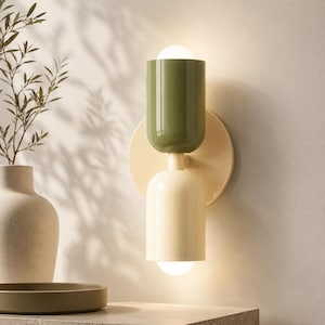 May include: A wall-mounted light fixture featuring a circular cream-colored backplate and two cylindrical shades. The top shade is olive green, and the bottom shade is cream, both emitting a soft glow from the exposed bulb ends.