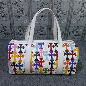 May include: White leather duffel bag with a colorful cross pattern. The bag features a zipper closure, two handles, and a decorative cross design in various colors, including red, blue, yellow, and black. The bag is approximately 12 inches long.