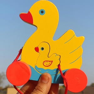 Könnte beinhalten: Ein gelbes Holz-Zugspielzeug in Form einer Ente mit einer kleineren Ente im Inneren. Die Ente hat einen roten Schnabel und orangefarbene Räder. Eine rote Schnur ist zum Ziehen befestigt. Spielzeug für Kinder.