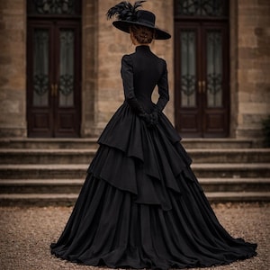 May include: A floor-length black gown with long sleeves and a high neckline, featuring a multi-layered, tiered skirt. The ensemble includes a wide-brimmed black hat adorned with dark feathers and matching black gloves, viewed from the back.