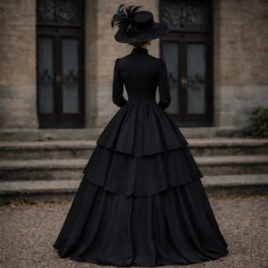 May include: A floor-length black dress with long sleeves, a high neckline, and a multi-tiered skirt. The garment is paired with a wide-brimmed black hat adorned with dark feathers, creating a cohesive monochromatic ensemble with a structured silhouette.