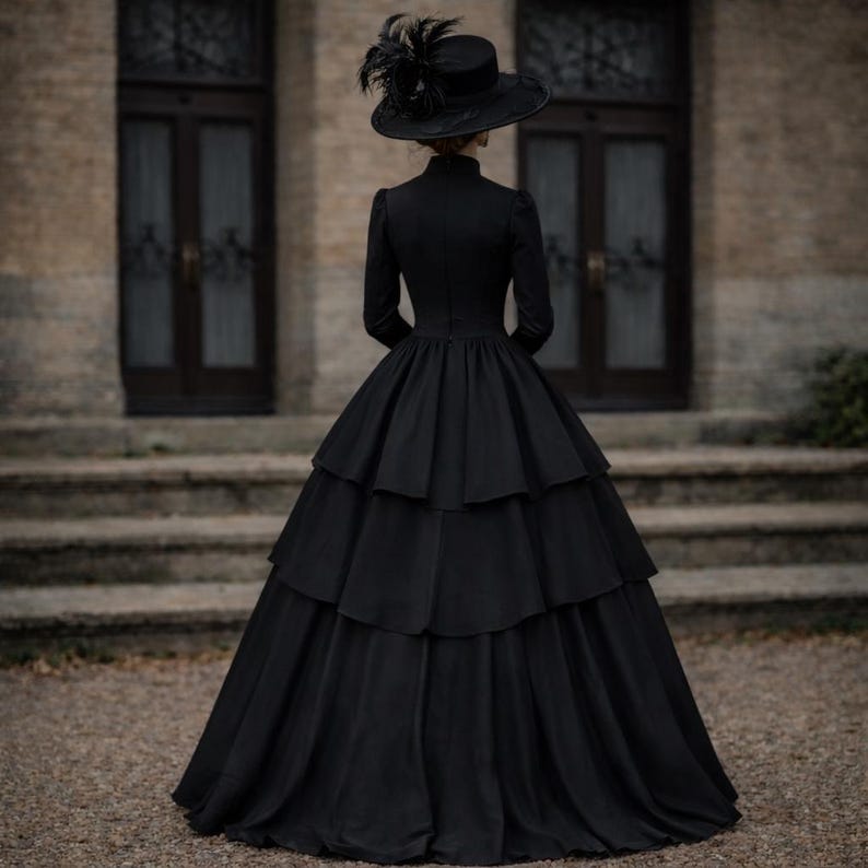 May include: A floor-length black dress with long sleeves, a high neckline, and a multi-tiered skirt. The garment is paired with a wide-brimmed black hat adorned with dark feathers, creating a cohesive monochromatic ensemble with a structured silhouette.